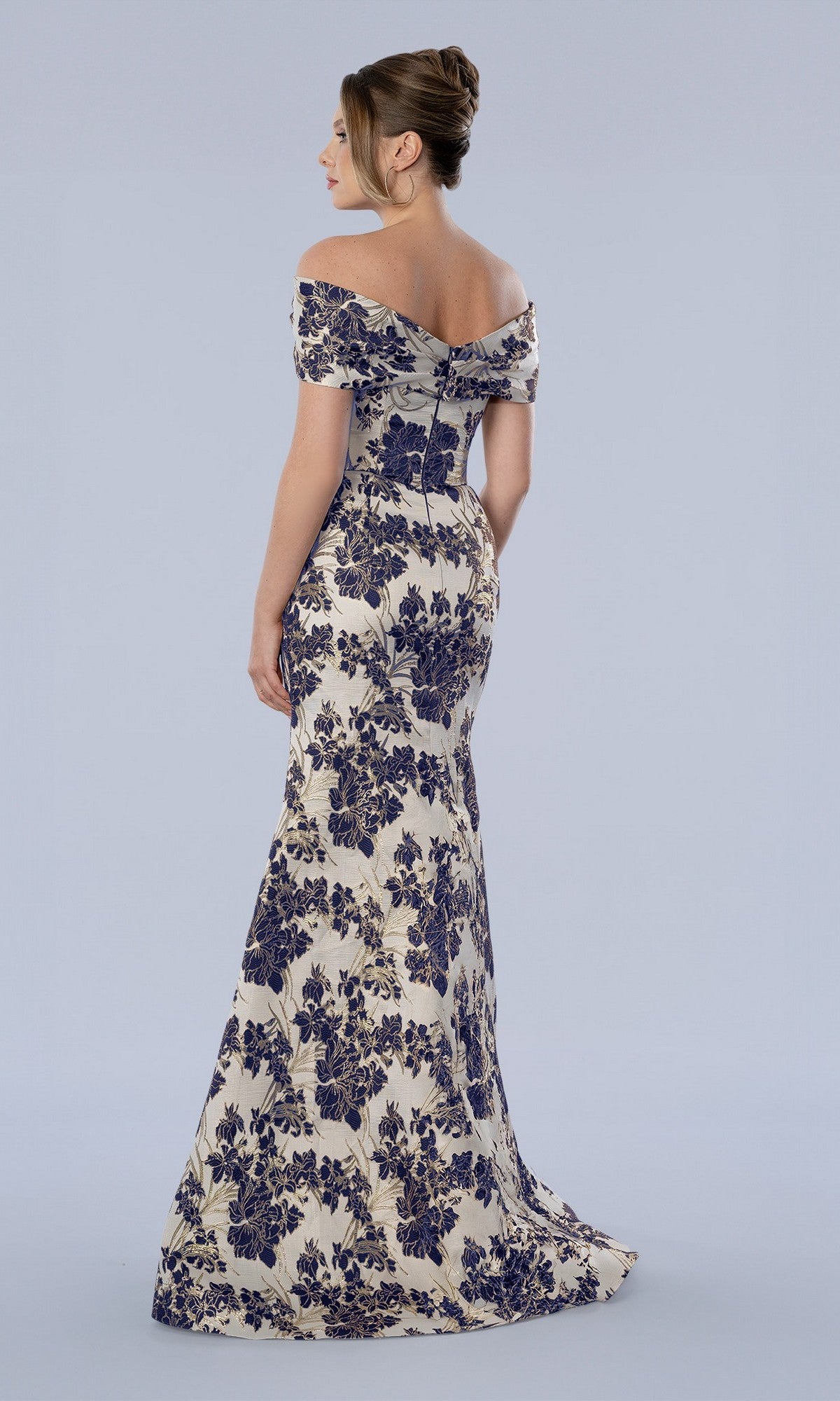 A woman in the Stella Couture 26359 long formal dress—an off-the-shoulder, floor-length gown featuring a floral brocade design—stands facing away against a plain light background, making it a beautiful option for a mother-of-the-bride dress.